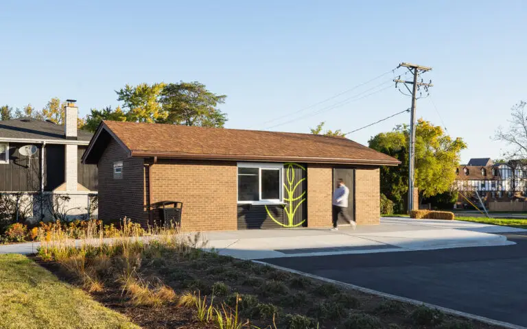 A small, single-story brick building is adaptively reused into a concession stand under a clear blue sky. The building has a low, brown shingled roof, one rectangular window, and a dark door painted with a bright green leaf graphic to reflect its location in the city of Hickory Hills for placemaking. A paved sidewalk and curb ramp run in front of the entrance, and landscaped plantings with grasses and shrubs line the foreground. One person is walking past the door, slightly blurred by motion. Utility poles, overhead wires, trees with early autumn foliage, and nearby houses appear in the background, placing the building within a quiet neighborhood setting.