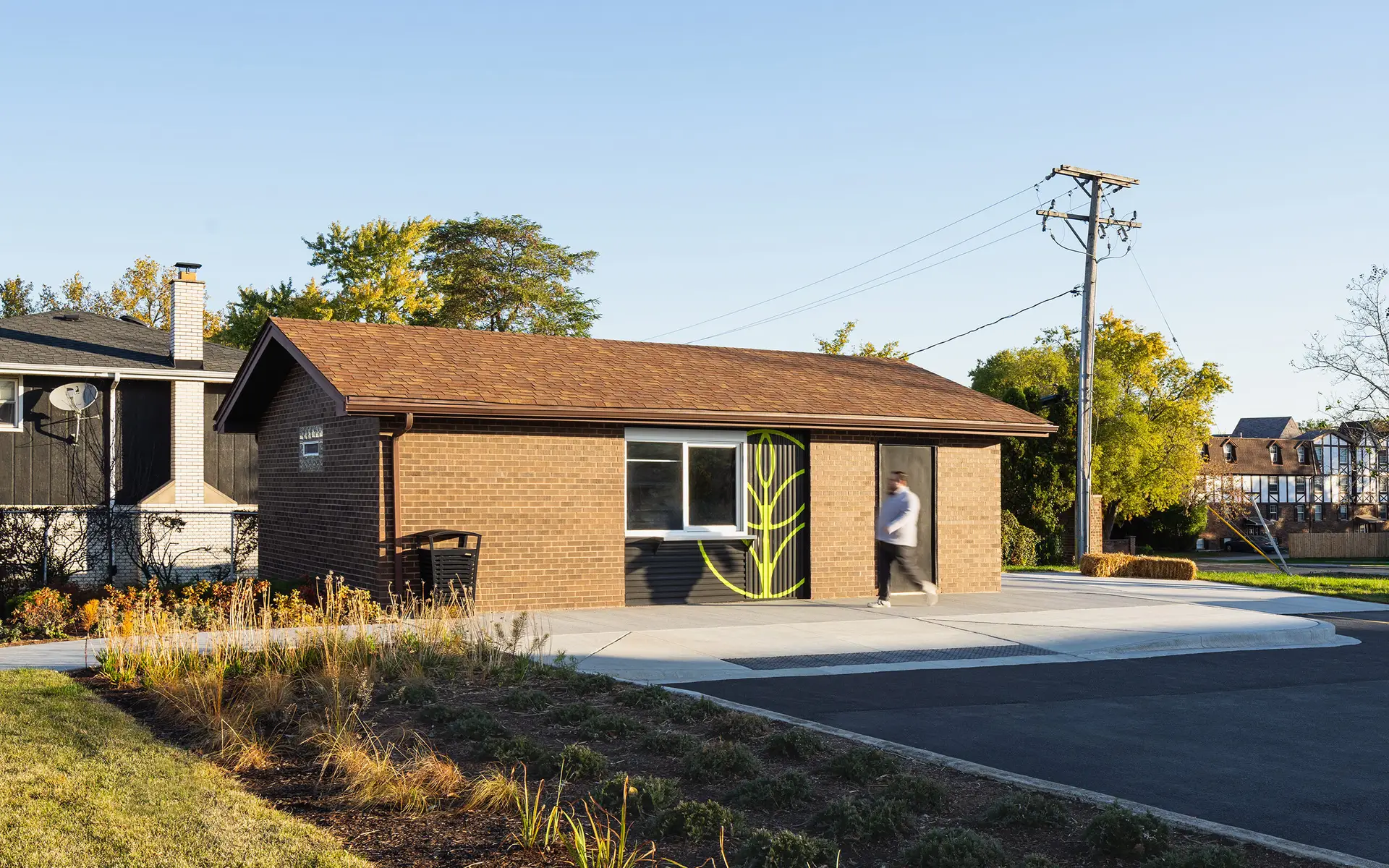 A small, single-story brick building is adaptively reused into a concession stand under a clear blue sky. The building has a low, brown shingled roof, one rectangular window, and a dark door painted with a bright green leaf graphic to reflect its location in the city of Hickory Hills for placemaking. A paved sidewalk and curb ramp run in front of the entrance, and landscaped plantings with grasses and shrubs line the foreground. One person is walking past the door, slightly blurred by motion. Utility poles, overhead wires, trees with early autumn foliage, and nearby houses appear in the background, placing the building within a quiet neighborhood setting.