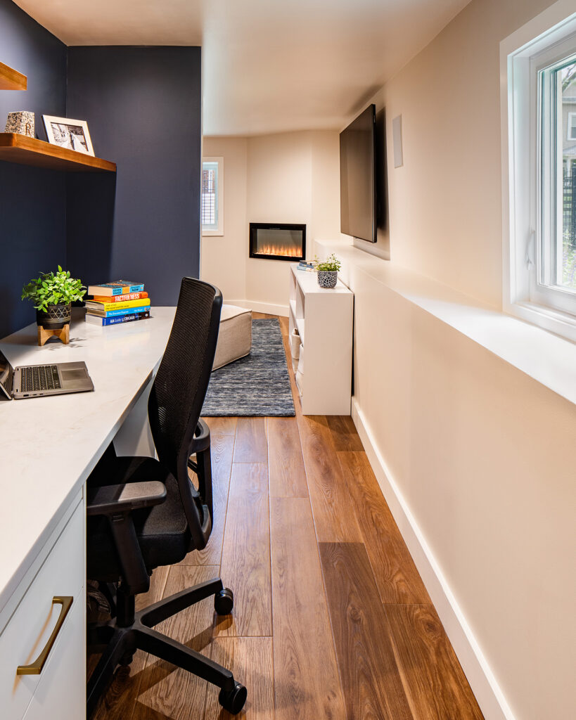 New office area in basement renovation consisting of built in millwork desk, navy blue walls, and quartz countertop, along with shelving and an adjacent window to the outside. Beyond you see the fireplace in the family room.